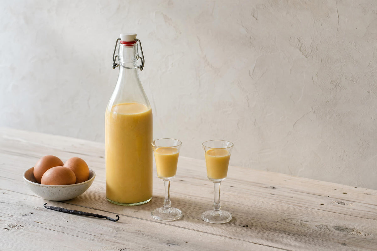 Selbst gemachter Eierlikör in einer Bügelflasche mit zwei gefüllten Likörgläsern, Eiern und Vanilleschote
